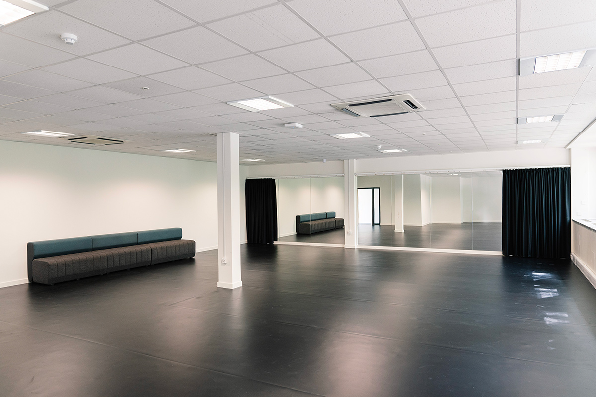 Internal view of the Development Hub studio, which is a bright room with a white ceiling and black floor. At the end of the room are floor to ceiling mirrors, which have black curtains at the end which can be covered. The space is empty, in order to rehearse.