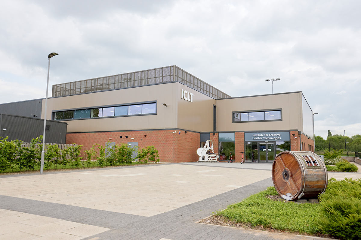 External view of the Institute Creative Leather Technologies building, which is a big building with brick on the lower level and smoother material at the top. Outside the door, where the sign of the building is, are 2 leather equipment's and green shrubbery.