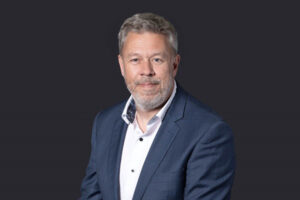 Roland Gray, Board of Governor at the University of Northampton. Roland is wearing a navy suit and the photo is taken in front of a dark board