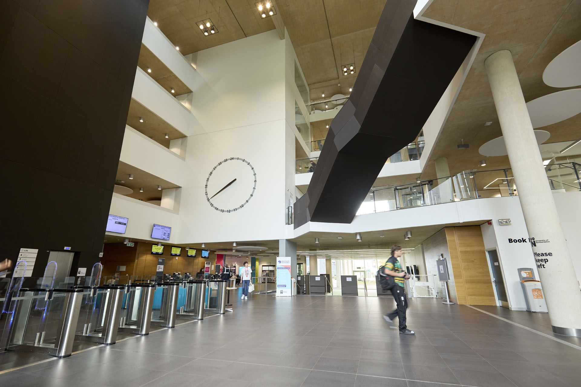 Internal view of the Learning Hub, showing barriers towards elevators and in the background the student information desk. There is a large clock on the wall. A person is walking through the spacious lobby.