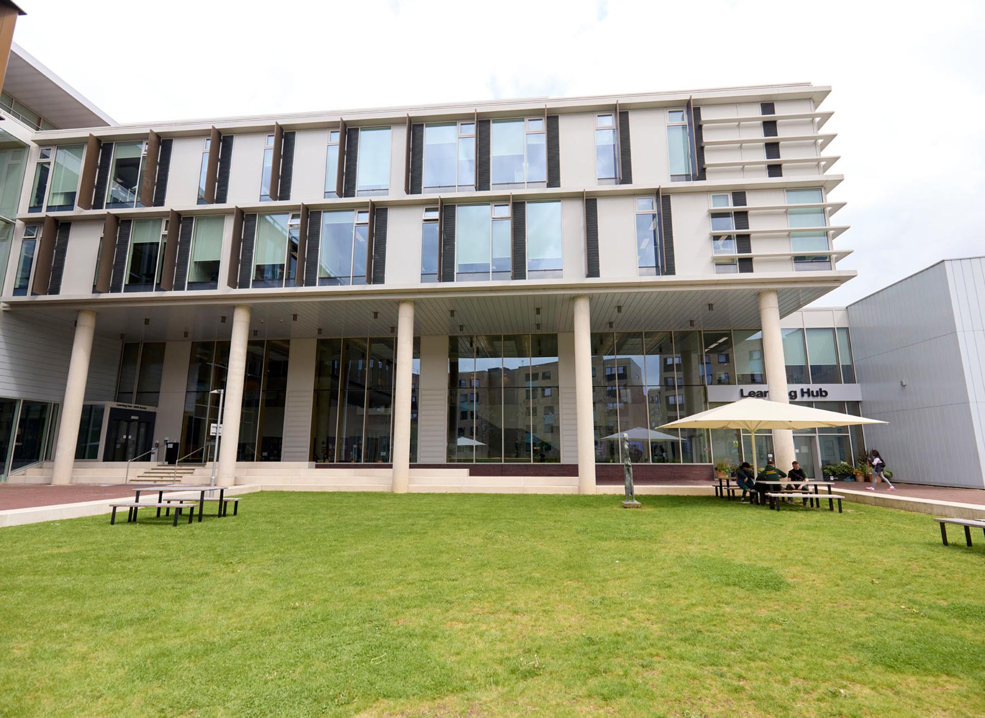 External view of the Learning Hub, with large windows and vertical pillars housing, adjacent to a green lawn with benches and an outdoor seating area with an umbrella.
