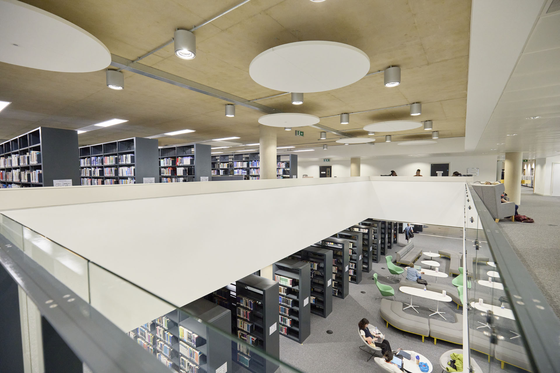 Two floors of the Learning Hub library, showing a spacious, modern interior with rows of bookshelves, a mezzanine level, and several students studying on couches and tables below.