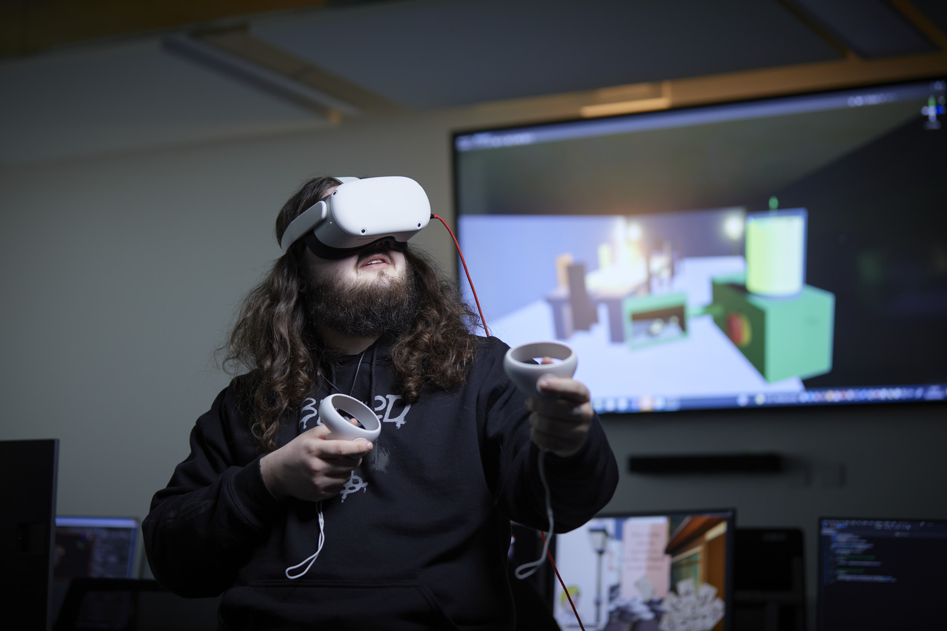 Student wearing a virtual reality headset, with the screen they are seeing behind them on a large screen on a wall. They have two devices in their hand.