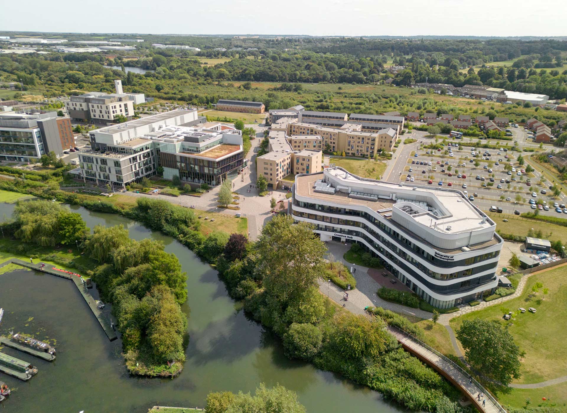 Aerial view of University of Northampton, Waterside campus featuring multiple modern buildings, a river, and surrounding greenery and open fields. Car parks and residential areas are visible in the background.