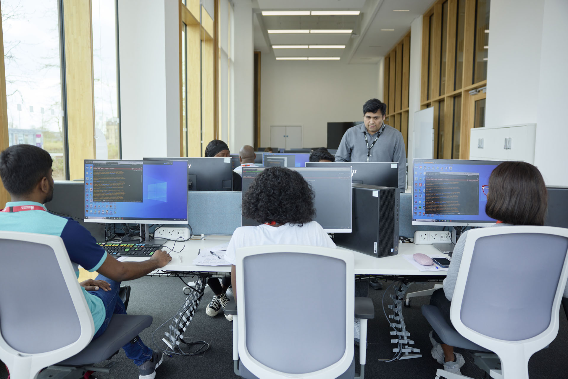 Students working at computer stations in a modern office with large windows. Behind them, an academic is standing while observing.