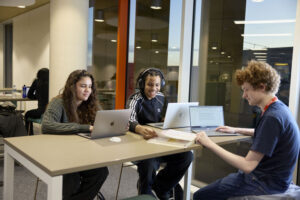 Three students sitting at a table with open laptops and a book in the Learning Hub. Two of the students are discussing a page in the open book.
