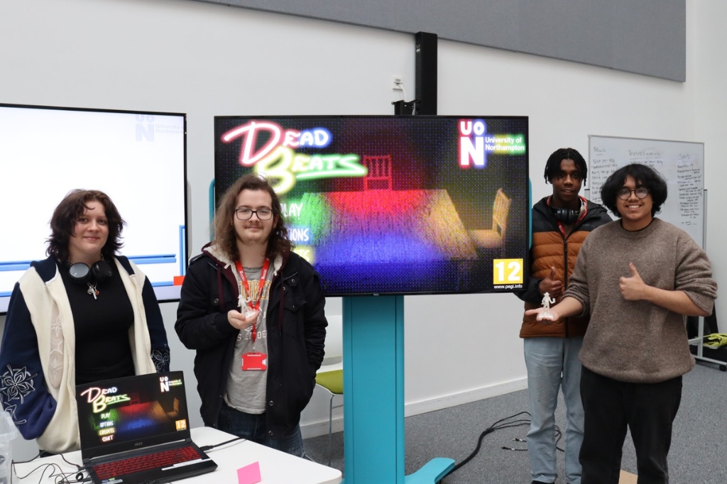 Group photo of three Games students in front of TV screen smiling at camera.