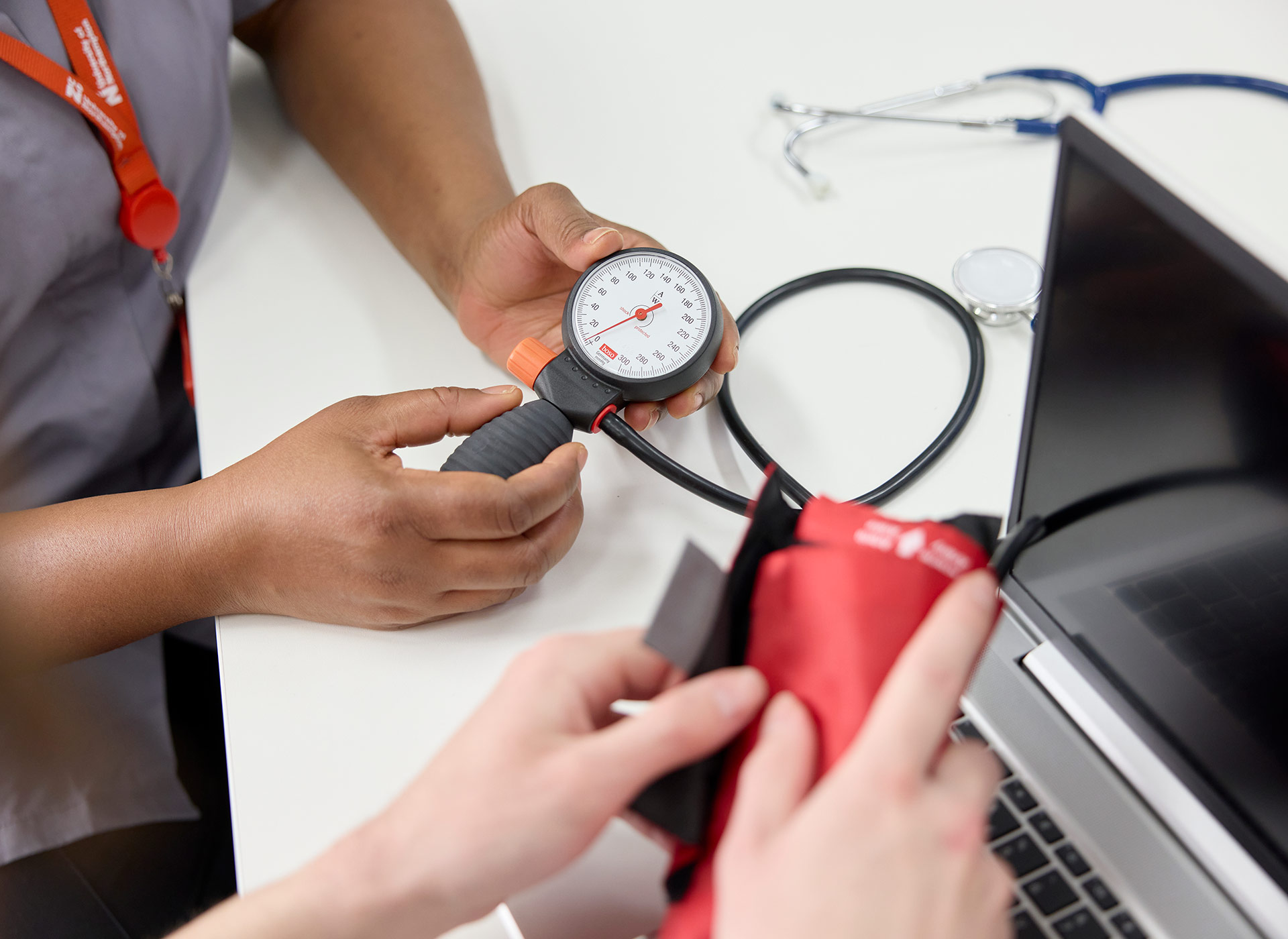 Two people examine a blood pressure monitor and cuff on a desk with a laptop and stethoscope.