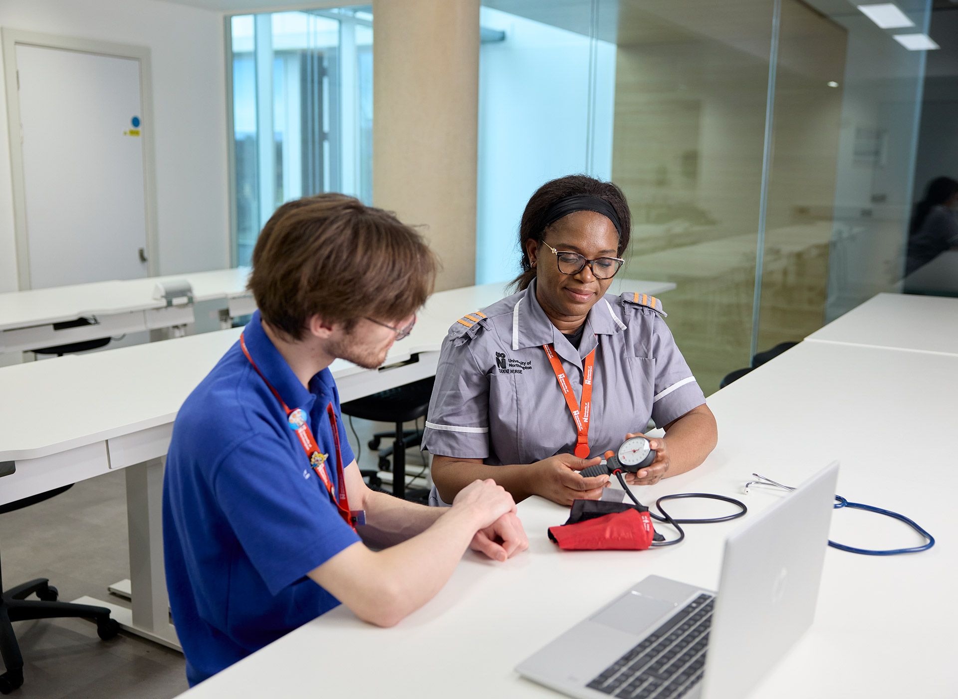 Two Learning Disability Students in a modern office setting, discussing a blood pressure monitor at a table with a laptop.