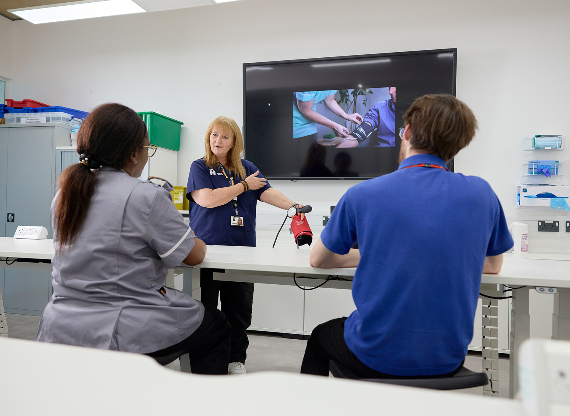 A Learning Disability academic is standing in front of a screen with an image of a nurse conducting a blood pressure test, while also holding out their own arm to demonstrate to two students sitting at white benches.