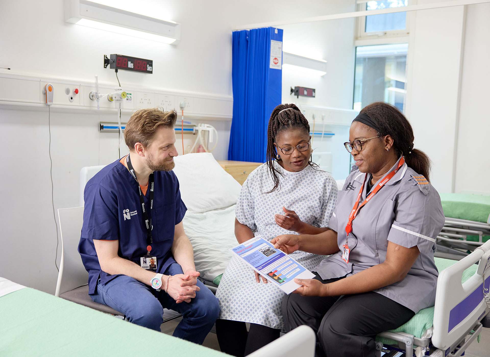 Two Learning Disability students discuss a brochure with a patient in a hospital room setting, on a ward.