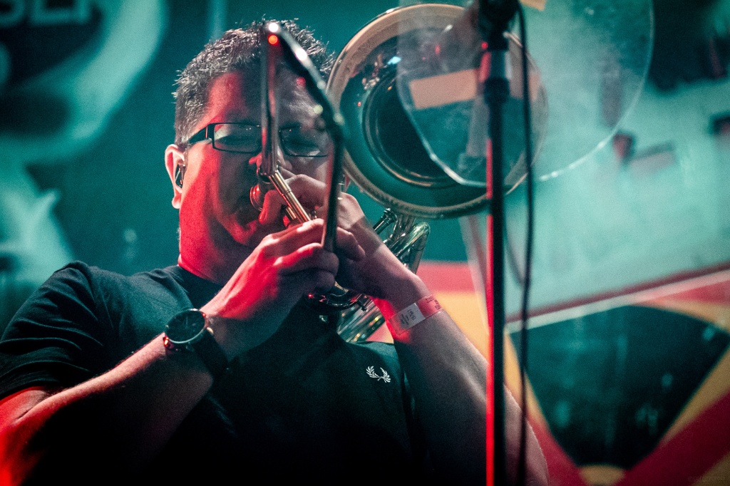A man plays a trombone passionately on stage, wearing glasses and a black shirt, with colorful lighting and a microphone stand in the foreground.