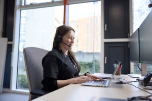 A female staff member wearing a headset sits at a desk, working on a laptop and looking at a large monitor in a bright office setting.