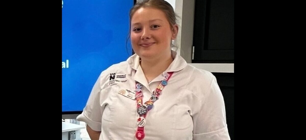Sofia Rose, Occupational Therapy graduate, wearing an UON uniform, smiles at the camera while standing next to a digital screen.