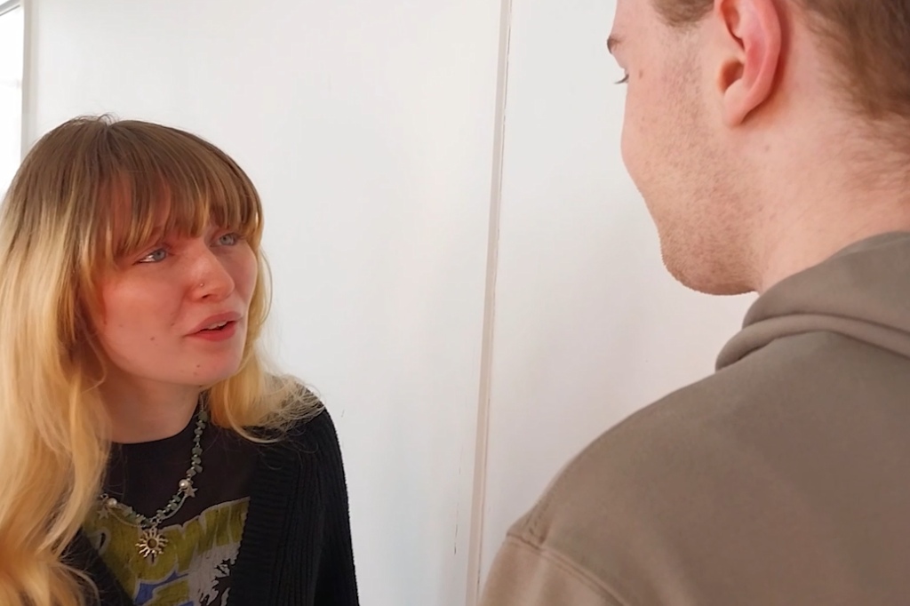 A woman with long blond hair and bangs speaks to a man facing away from the camera in a brightly lit room.
