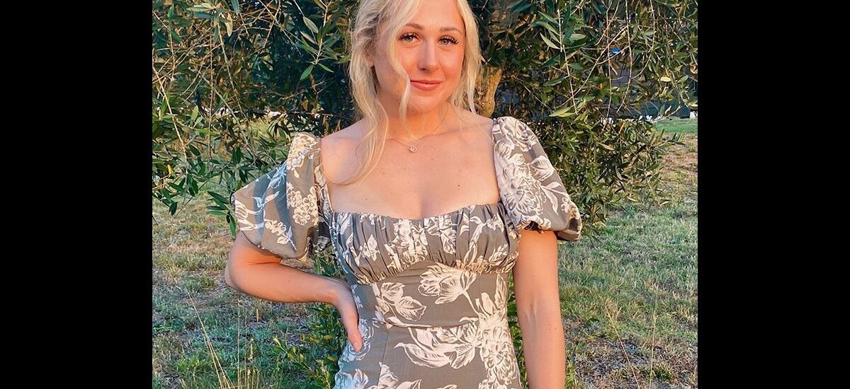 A woman with blonde hair in a loose updo stands outdoors, wearing a gray floral dress with puffed sleeves. Shrubs and trees are in the background.