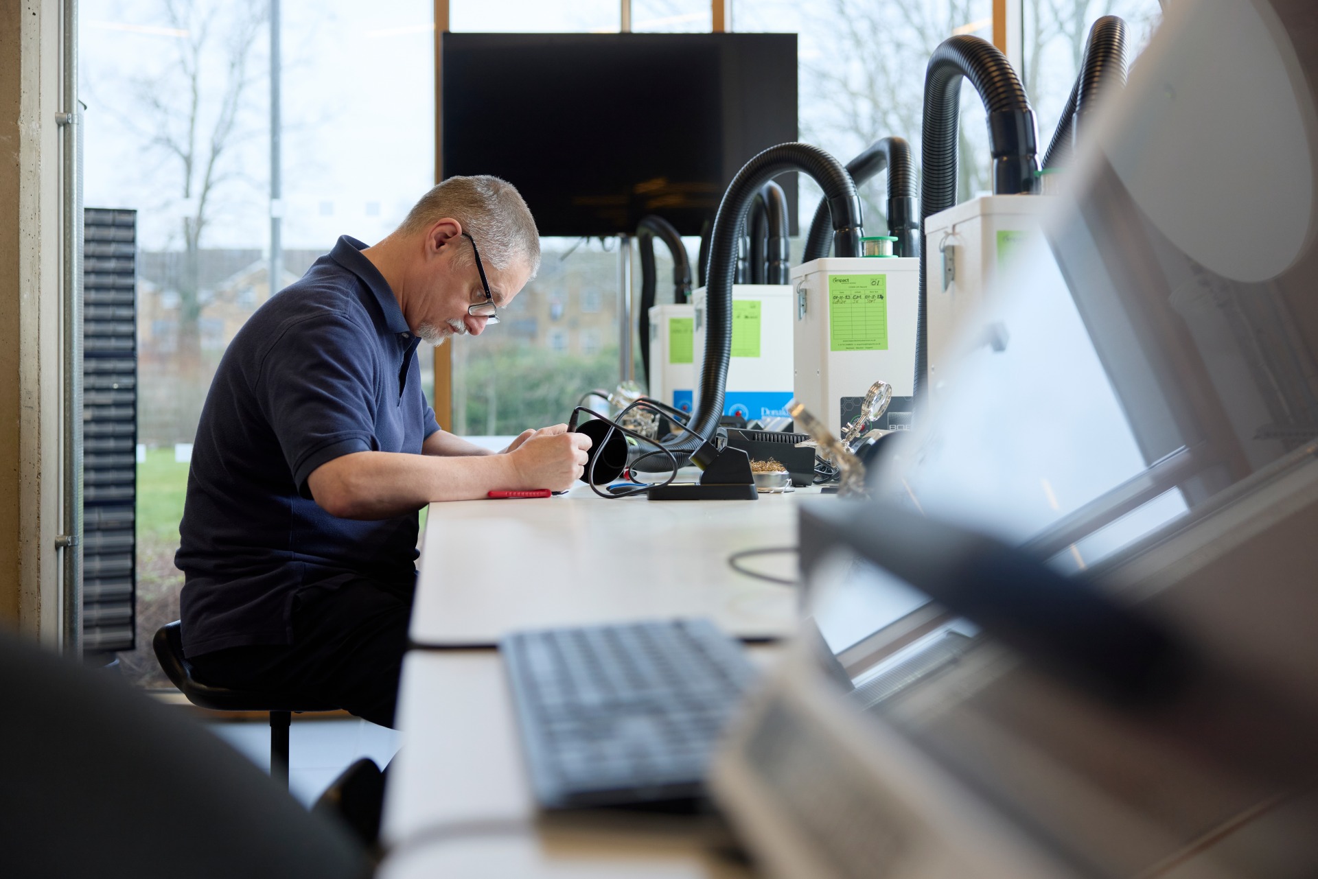 A man sits at a desk in a laboratory or workshop, working with electronic equipment and tools, with computers and devices visible on the workstations.