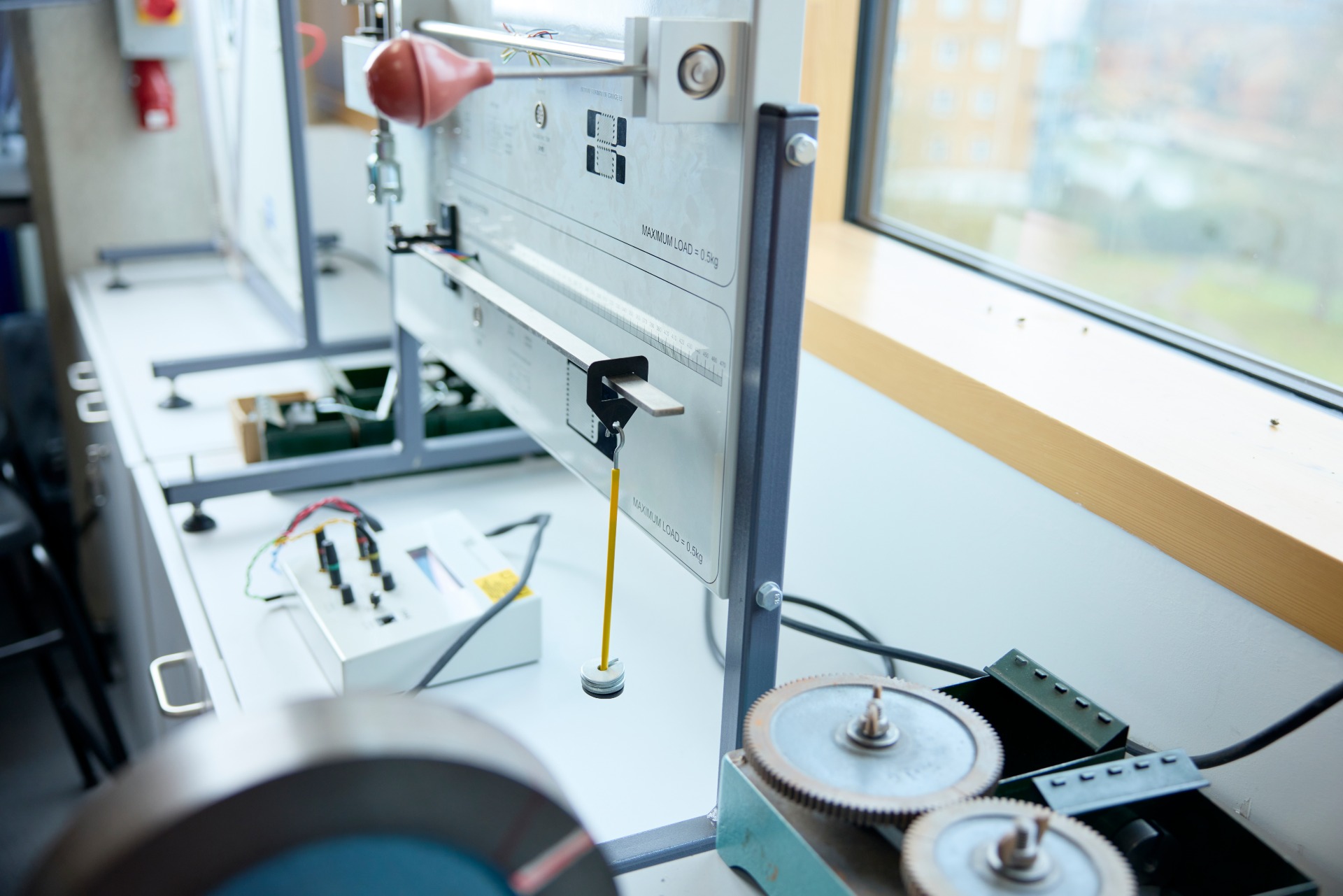 A close-up of a laboratory apparatus with gears, wires, and measuring devices on a white table next to a window.