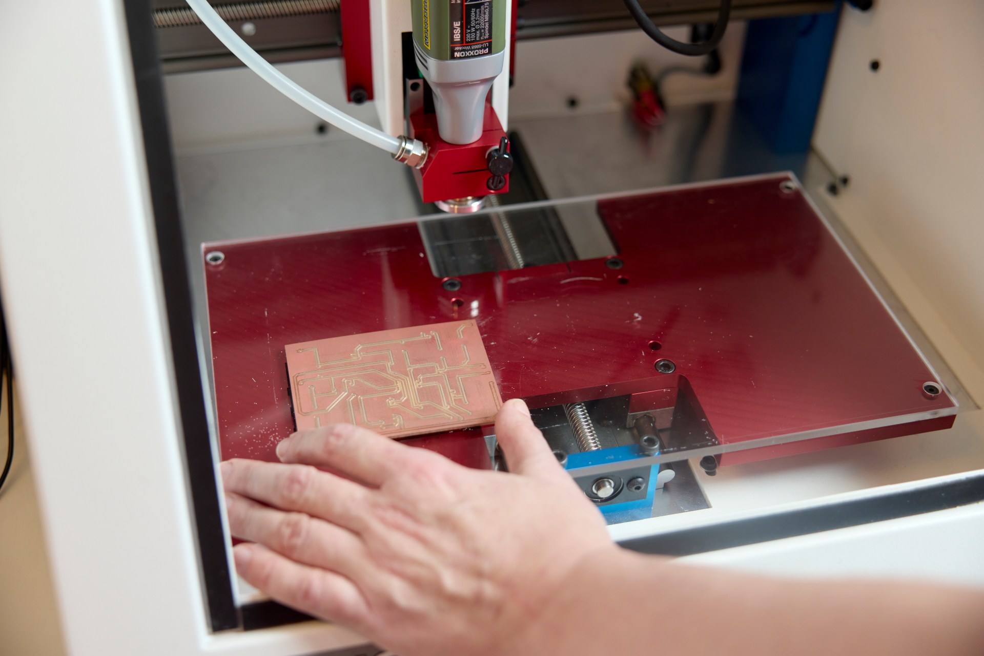 A hand operates a CNC milling machine engraving patterns onto a copper circuit board inside a protective enclosure.