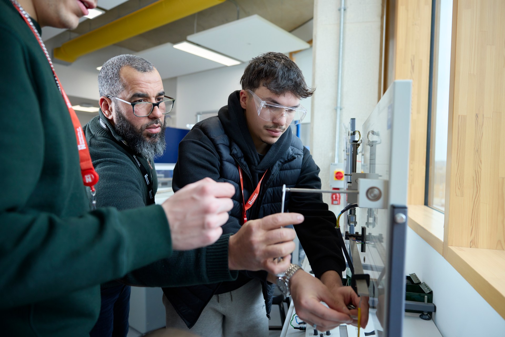 Three people work together in a laboratory, with one person supervising as the others use lab equipment and wear safety goggles.