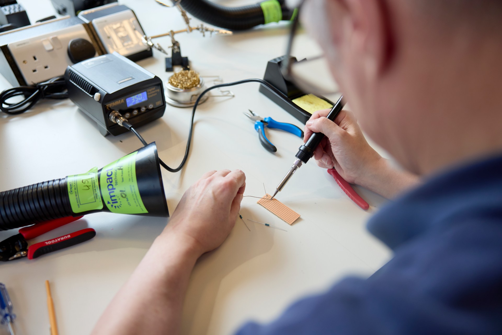 A person uses a soldering iron to assemble electronic components on a circuit board at a workbench with various tools and equipment.