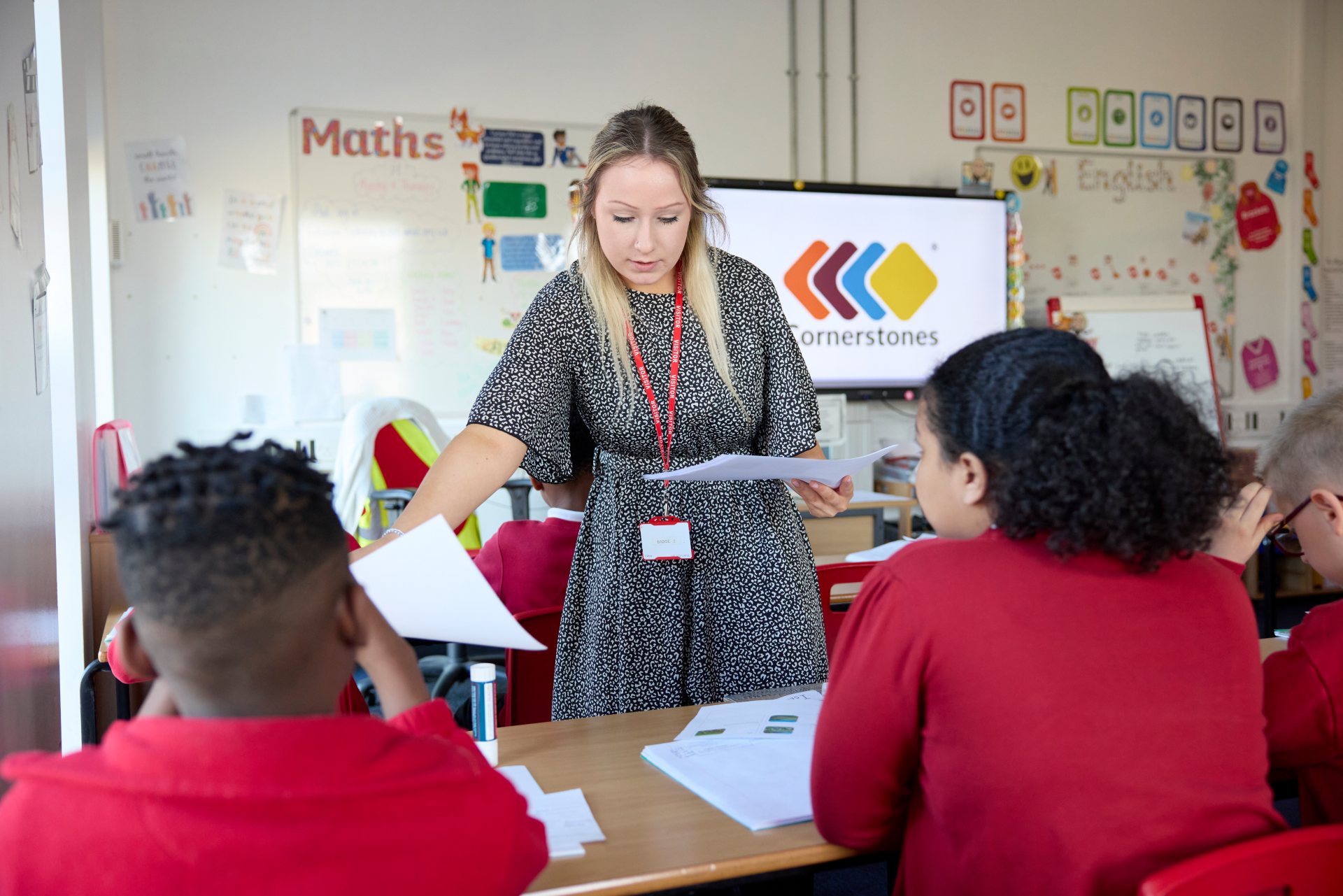 A teacher hands out papers to students in a classroom, with a screen displaying the Cornerstones logo in the background. Several students in red uniforms are seated at desks.