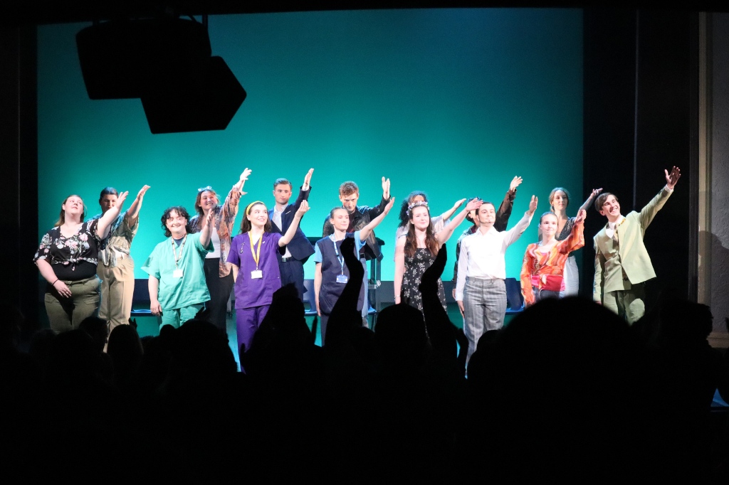 A group of performers stands in a row on stage, raising one arm, facing an audience with silhouetted heads visible in the foreground.