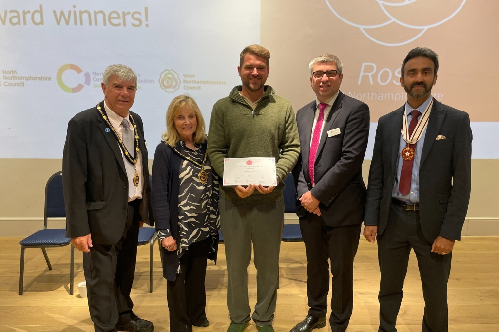 Five people stand indoors, one holding a certificate. All are dressed formally except the certificate-holder, who is in casual clothing. A presentation screen and chairs are visible in the background.