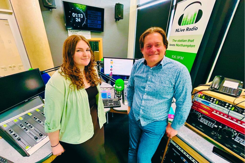 Two people stand in a radio studio with control panels and microphones, next to an NLIVE Radio banner. Computer monitors and studio equipment are visible in the background.