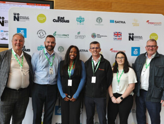 Six visitors to the UON Sustainability Summit 2023, standing in front of the board with sponsor logos on them. They are all wearing green lanyards and smiling.