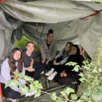 Five University students sit inside a makeshift tent in a forest, surrounded by greenery.